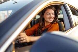 Beautiful young happy smiling woman driving her new car at sunset. Portrait of smiling young Caucasian woman sitting alone on driver seat with fasten belt while driving modern car.